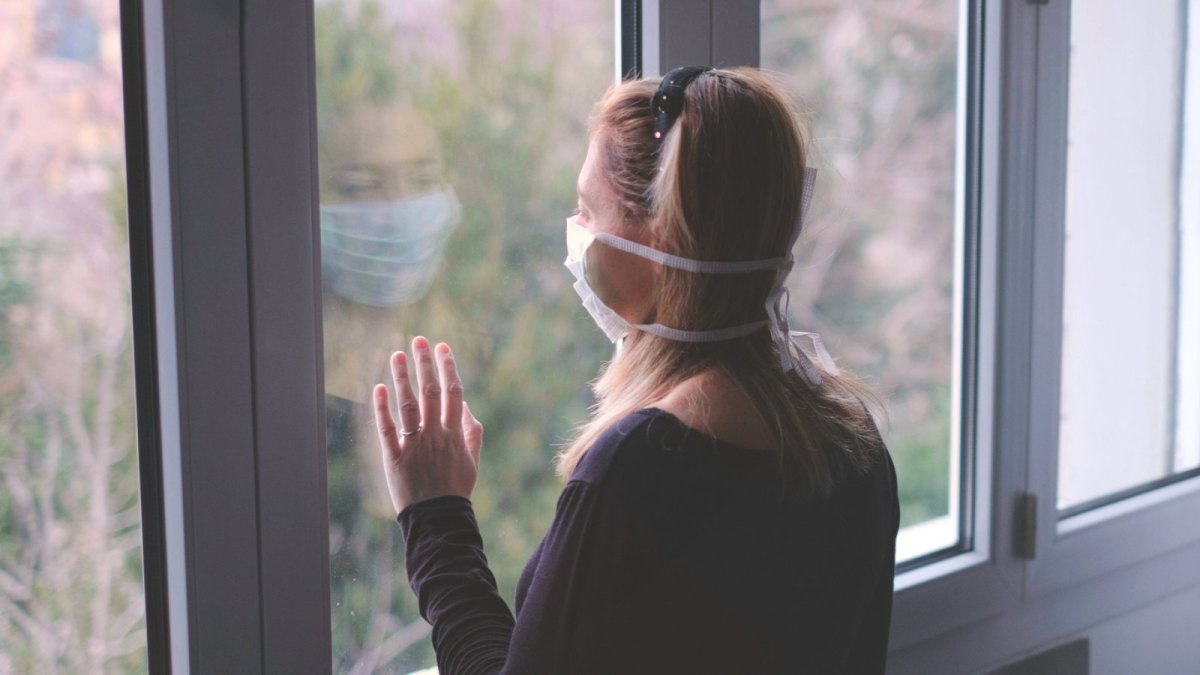 Frau mit Mundschutz schaut aus dem Fenster.