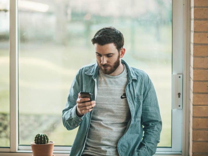 Mann steht vor einem Fenster und schaut auf sein Smartphone.