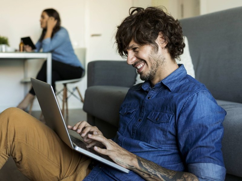 Ein Mann sitzt vor einer Couch mit seinem Laptop und lehnt sich an die Sitzfläche an. Im Hintergrund sitzen drei Personen an einem Tisch.