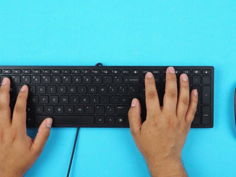 Eine schwarze Tastatur und eine schwarze PC-Maus auf einem hellblauen Untergrund. Zwei Hände tippen auf der PC-Tastatur.