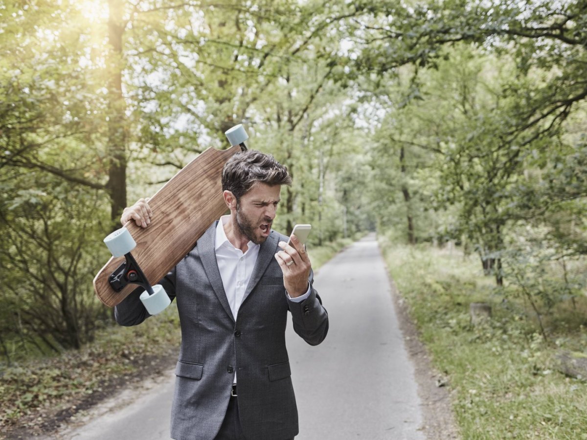 Ein Mann läuft mit einem Longboard durch den Wald und schreit sein Smartphone an.