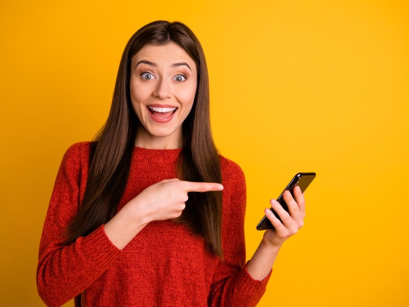 Frau zeigt frühlich mit dem Finger auf ihr Handy.