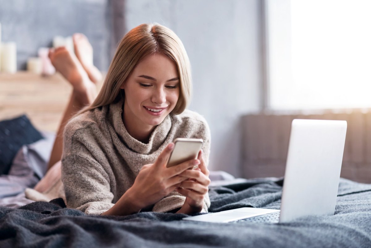 Frau liegt mit einem Handy vor dem Laptop.