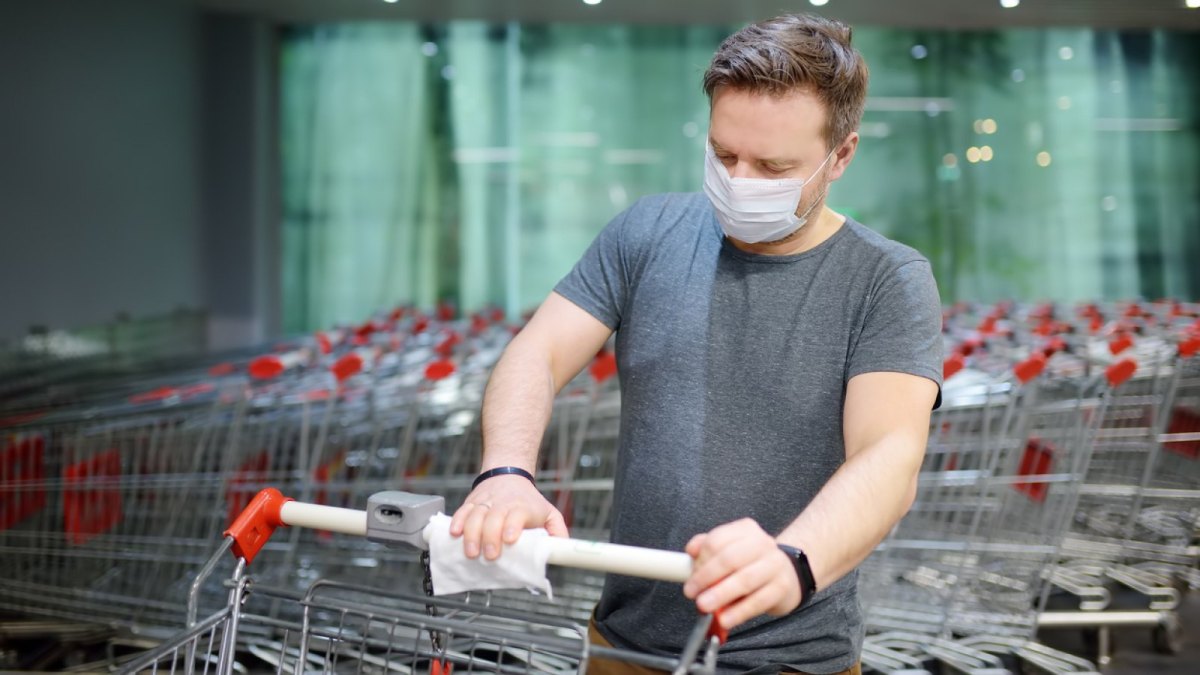 Mann mit Maske desinfiziert einen Einkaufswagen.