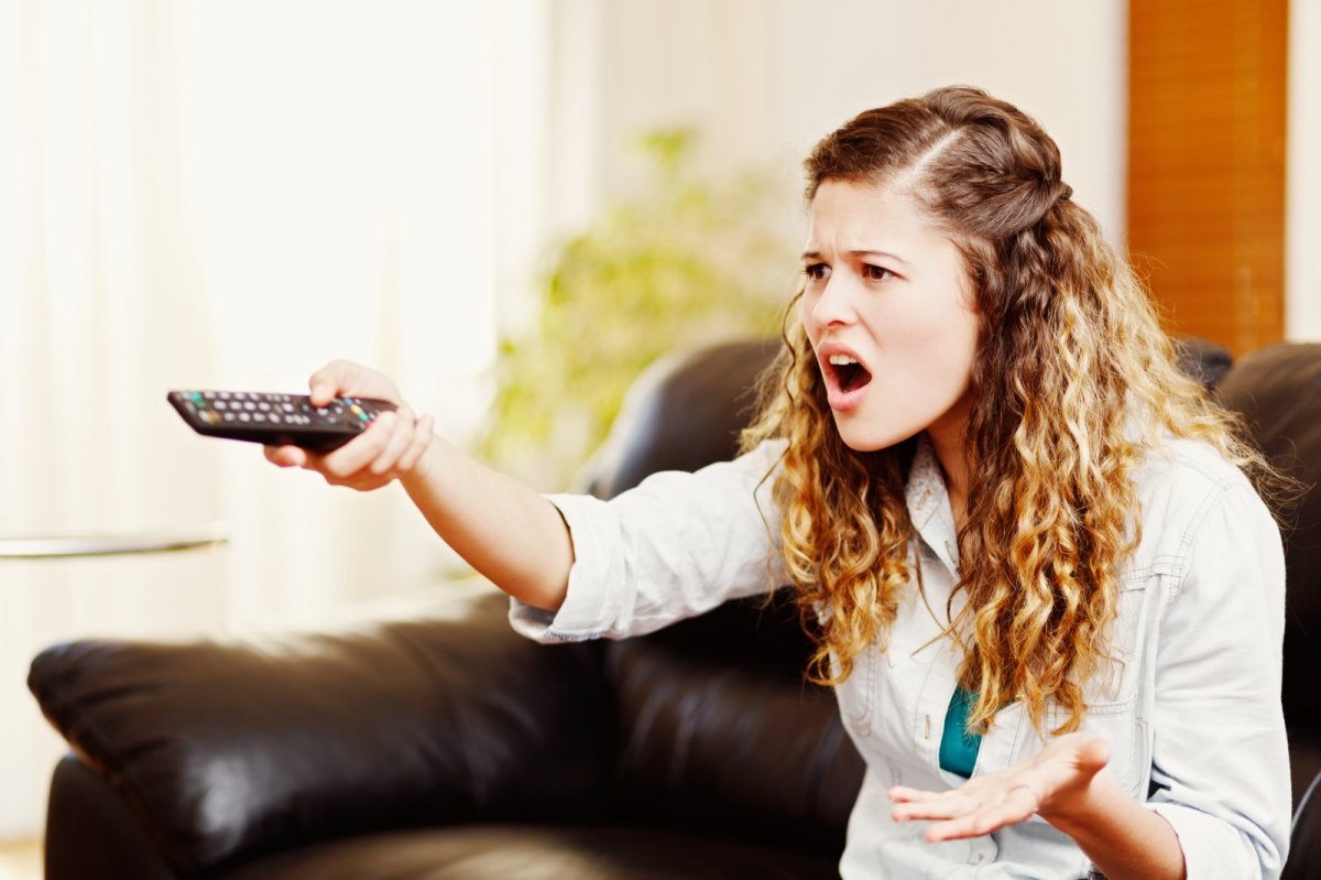 Frau zielt mit einer Fernbedienung ärgerlich auf einen Fernseher.