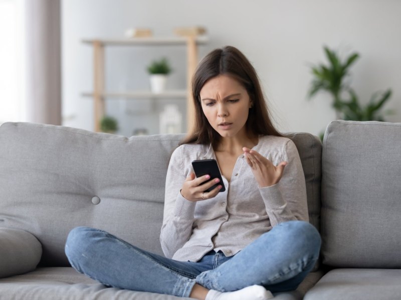 Frau sitzt mit ihrem Handy auf dem Sofa und schaut genervt auf das Gerät.