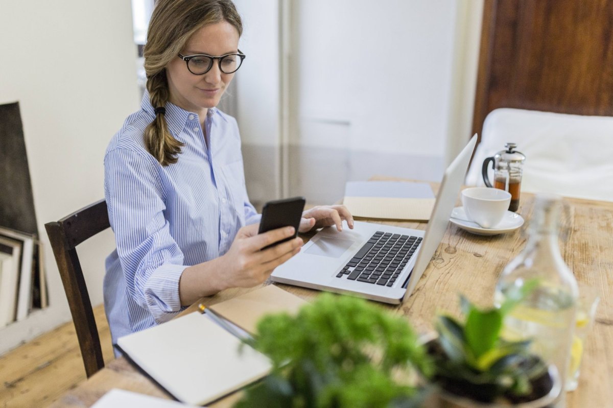 Frau sitzt am Laptop und schaut auf ihr Smartphone