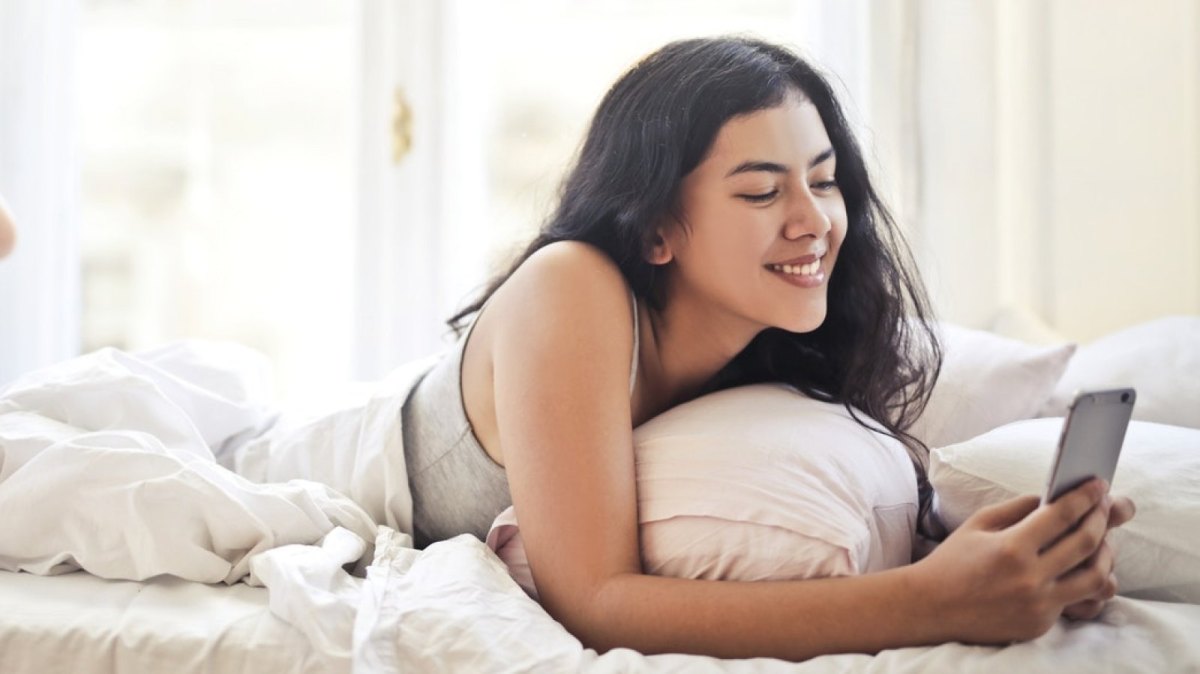 Eine Frau liegt im Bett und schaut lächelnd auf ihr Smartphone.