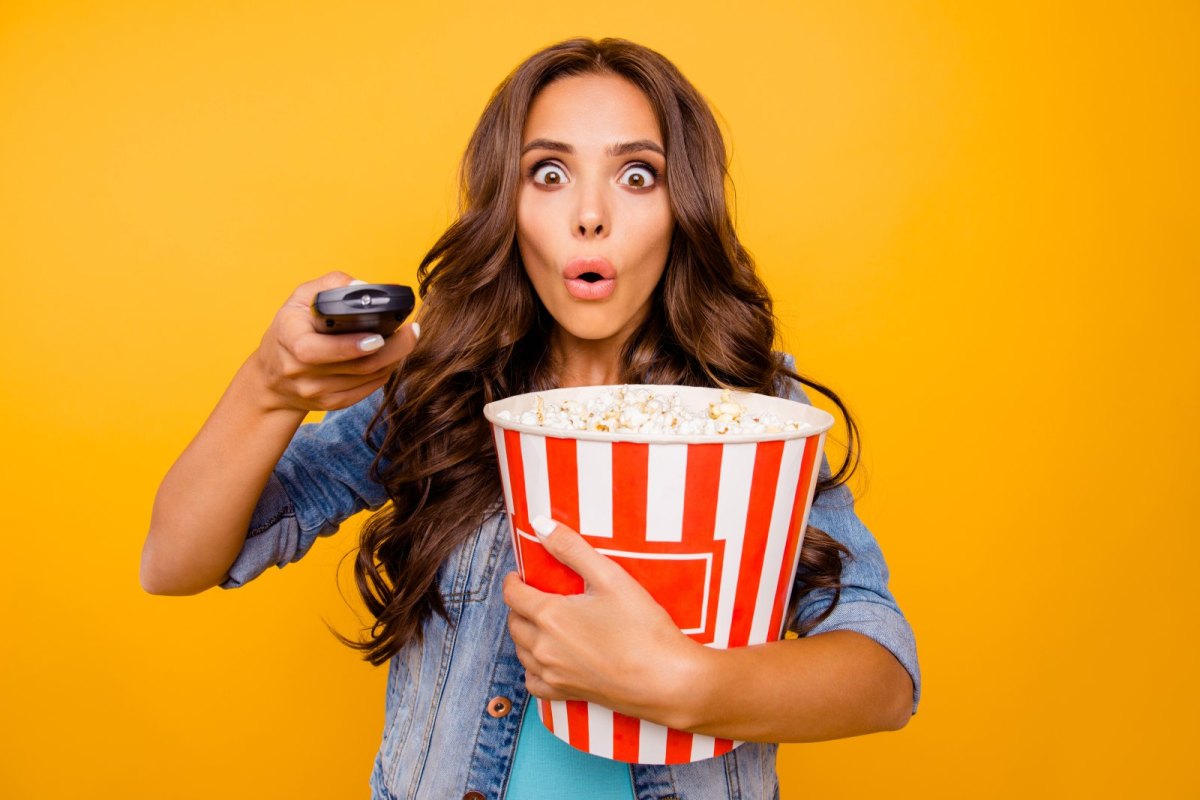 Frau mit Popcorn in der Hand drückt mit erstauntem Gesichtsausdruck auf eine Fernbedienung.