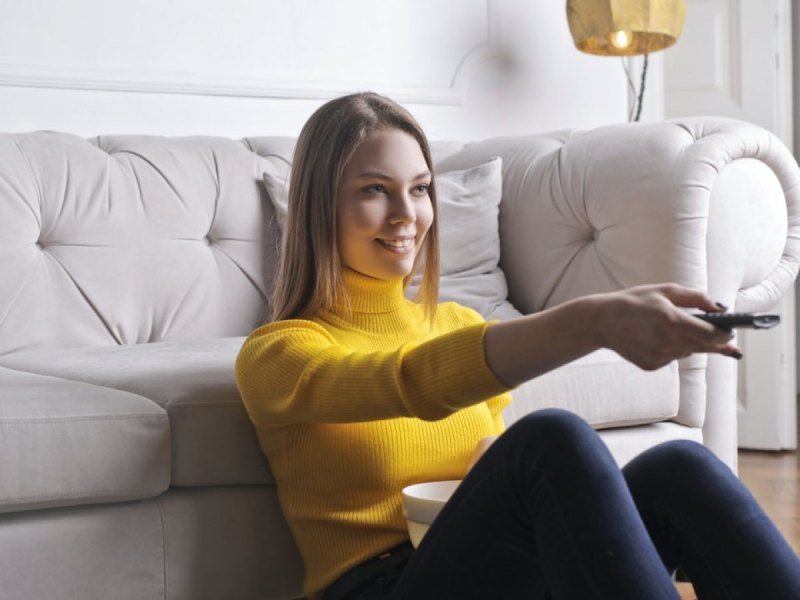 Eine Frau sitzt vor einem Sofa und hat eine Fernbedienung in der Hand.