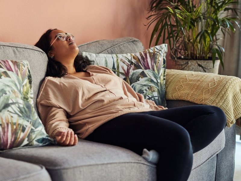 Frau sitzt nachdenklich und geschafft auf dem Sofa und blickt nach oben.