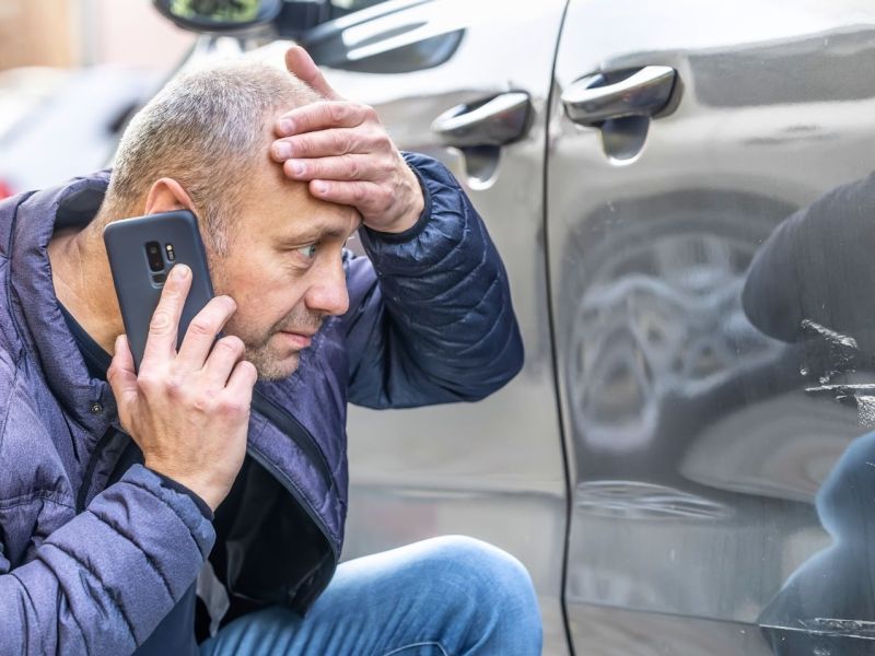 Mann telefoniert während er schockiert auf einen Kratzer an einem Auto schaut.