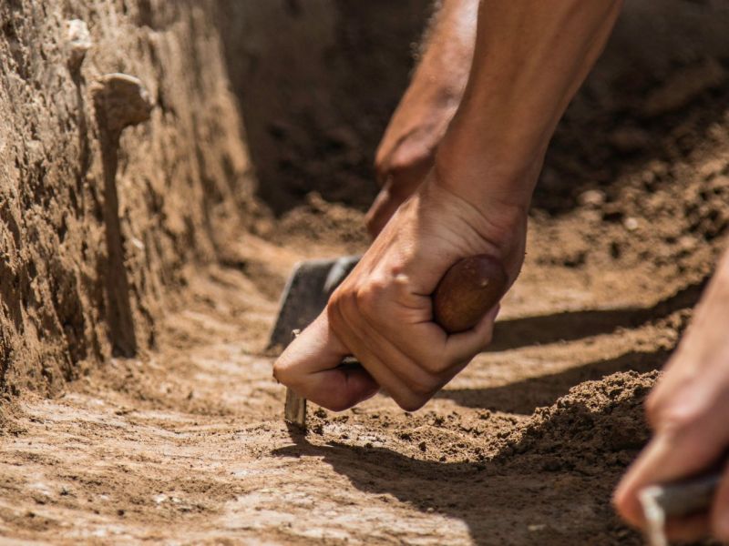 Hände graben mit Werkzeugen an einem archäologischen Fund.