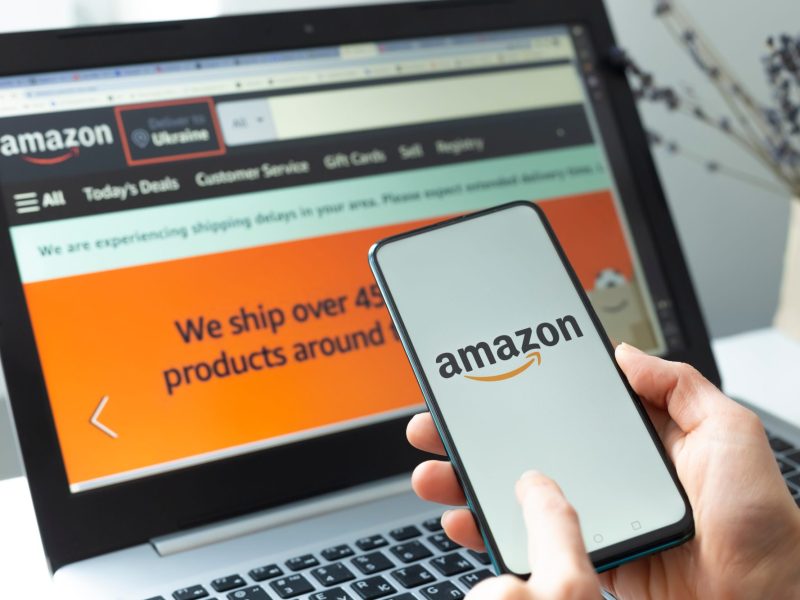 Amazon Logo auf einem Smartphone, im Hintergrund ist die Amazon-Website auf einem Laptop offen.