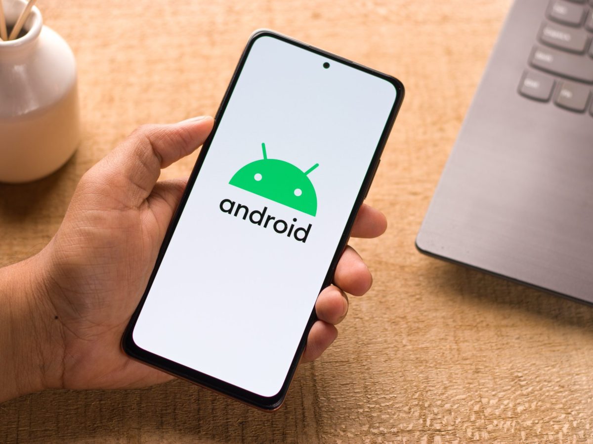 Eine Person hält ein Handy in der Hand. Auf dem Display ist das Android-Logo zu sehen. Im Hintergrund befindet sich ein Laptop auf einem Tisch.