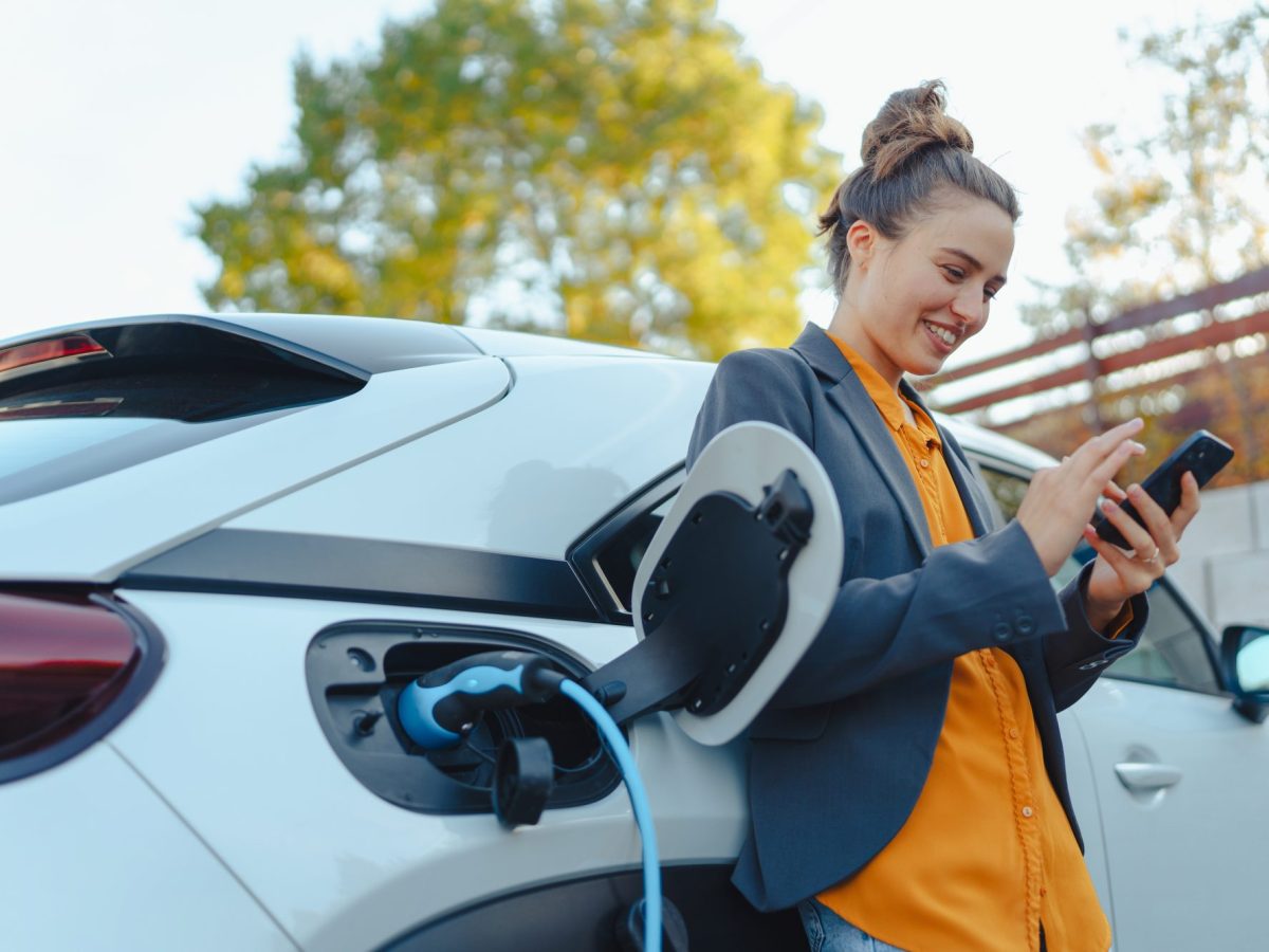 Eine Frau steht mit Smartphone an Elektroauto gelehnt, das währenddessen geladen wird.