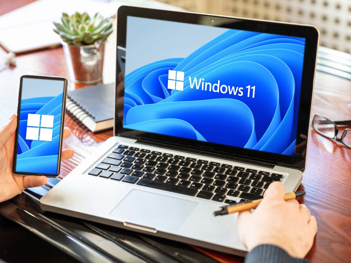 Auf einem Laptop wird das Windows 11-Logo angezeigt. Daneben hält eine Person ein Handy in der Hand. Auf dem Display wird das Windows-Logo angezeigt.