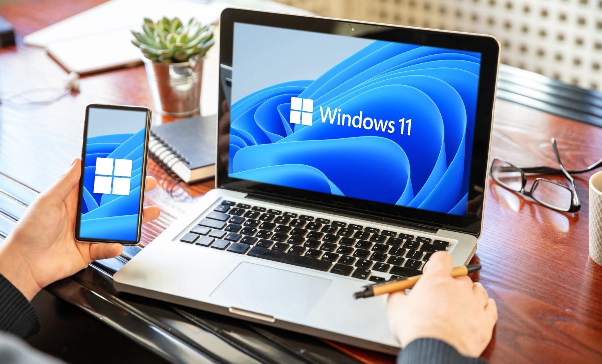 Auf einem Laptop wird das Windows 11-Logo angezeigt. Daneben hält eine Person ein Handy in der Hand. Auf dem Display wird das Windows-Logo angezeigt.