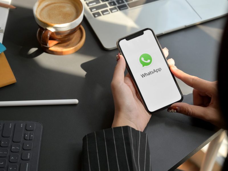 Eine Person hält ein Handy in der Hand, auf dessen Display ist das WhatsApp-Symbol zu sehen.
