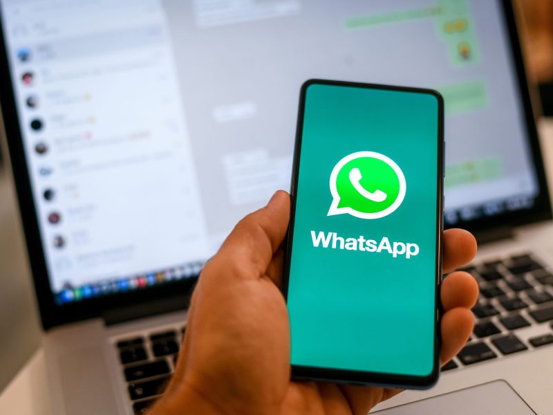 Person hÃ¤lt Handy in der Hand. Auf dem Bildschirm ist das WhatsApp-Logo zu sehen, im Hintergrund steht ein Laptop, auf dem WhatsApp geÃ¶ffnet ist.