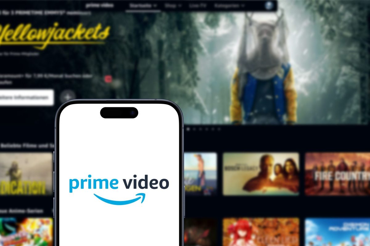 Auf einem Handy-Display wird das Amazon Prime Video-Logo angezeigt. Im Hintergrund ist ein Fernseher zu sehen, auf dem mehrere Video-Inhalte angezeigt.