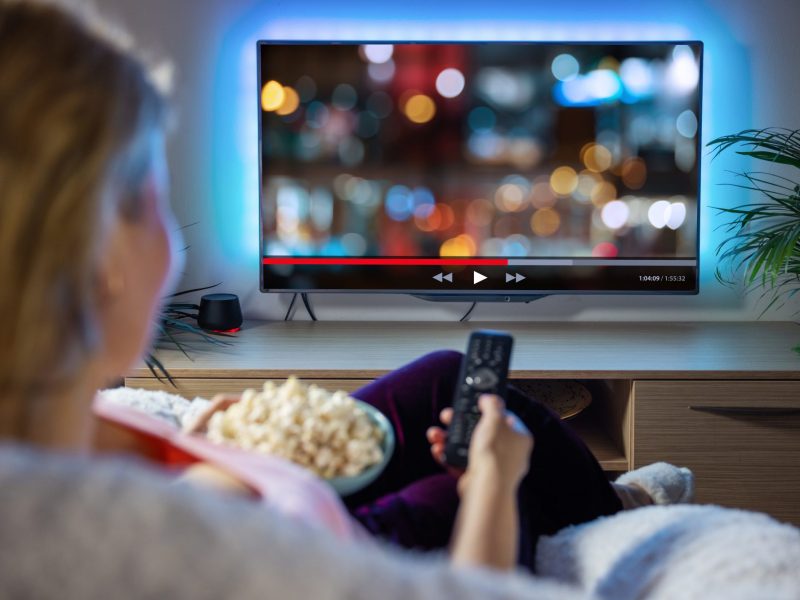 Frau sitzt mit Fernbedienung in der Hand und Popcorn auf dem Schoß vor einem Fernseher.