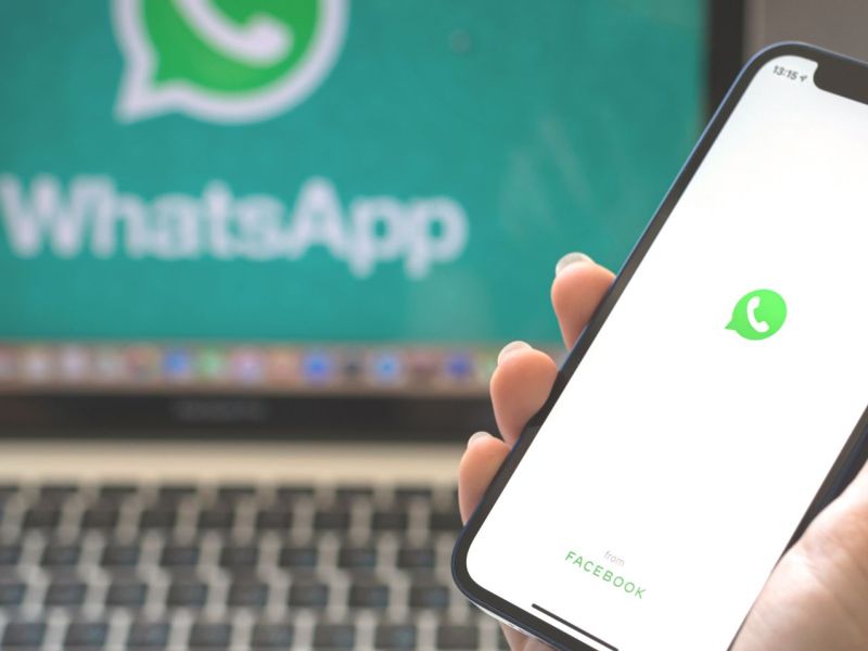 Hand hält Smartphone mit Whatsapp-Symbol vor Laptop mit demselben Symbolen.